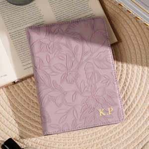 May include: A lavender passport holder with an embossed floral design and gold initials "K.P." is displayed on a woven surface. The passport holder is next to an open book and a perfume bottle.