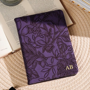 May include: A purple passport holder with a floral design and gold initials "AB". The cover is embossed with a detailed botanical pattern. A candle, a perfume bottle, and an open book are in the background.