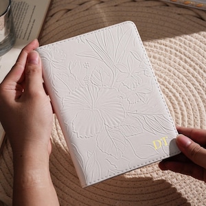 May include: White passport holder with embossed floral design. The cover features a detailed pattern of flowers and leaves. The initials "DT" are in gold on the lower right corner. The passport holder is held by a person.