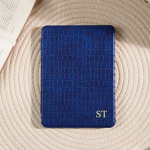 Pode incluir: Uma capa retangular azul cobalto com um padrão texturizado semelhante à pele de crocodilo. As iniciais "ST" estão em letras douradas. A capa está sobre uma superfície bege tecida.