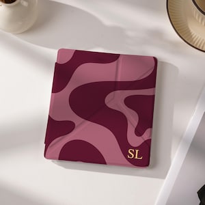 May include: A square protective case for an electronic device featuring a wavy, abstract pattern in shades of dark burgundy and dusty rose. The bottom right corner displays the gold-coloured initials SL in a serif font on the smooth surface.