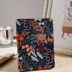 以下が含まれることがあります： オレンジ、ピーチ、白、緑の花柄が刺繍された濃紺のノート。表紙には、さまざまな花や葉の細かなステッチが施されています。ノートは長方形で、閉じられているように見えます。