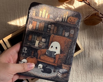 Könnte beinhalten: Eine dekorative Tablet-Hülle mit einer Halloween-Illustration. Sie zeigt einen Geist, der in einer Bibliothek mit Bücherregalen, Kerzen, Kürbissen und einer schwarzen Katze liest. Die Hülle wird von einer Hand gehalten.