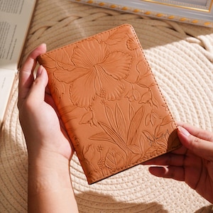 May include: A tan leather journal with an embossed floral design featuring a large hibiscus flower and leaves. The journal is held open, revealing the intricate details of the embossed pattern. The journal is a beautiful accessory.
