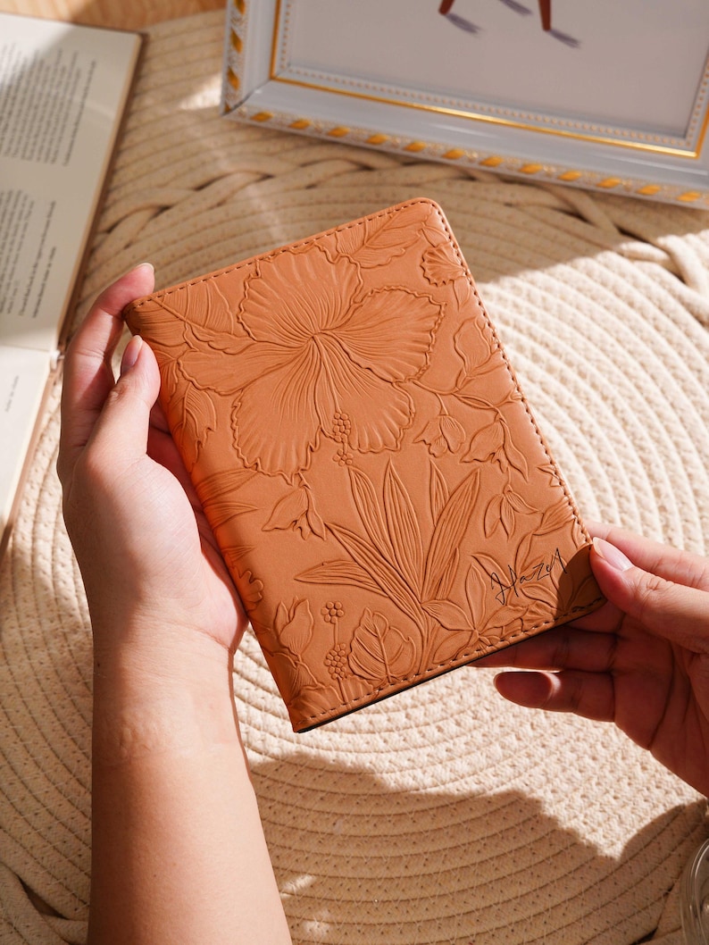 May include: A tan leather journal with an embossed floral design featuring a large hibiscus flower and leaves. The journal is held open, revealing the intricate details of the embossed pattern. The journal is a beautiful accessory.