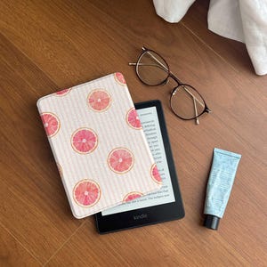 May include: A black Kindle e-reader with a protective cover featuring a light pink and white vertical striped pattern with pink grapefruit slices. Beside the device are thin-rimmed brown glasses and a small blue tube of hand cream.
