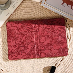 May include: A red floral-patterned clutch with a detailed embossed design. The clutch is rectangular and features a variety of flowers and leaves. The image is set on a straw mat with a candle, a book, and a perfume bottle.