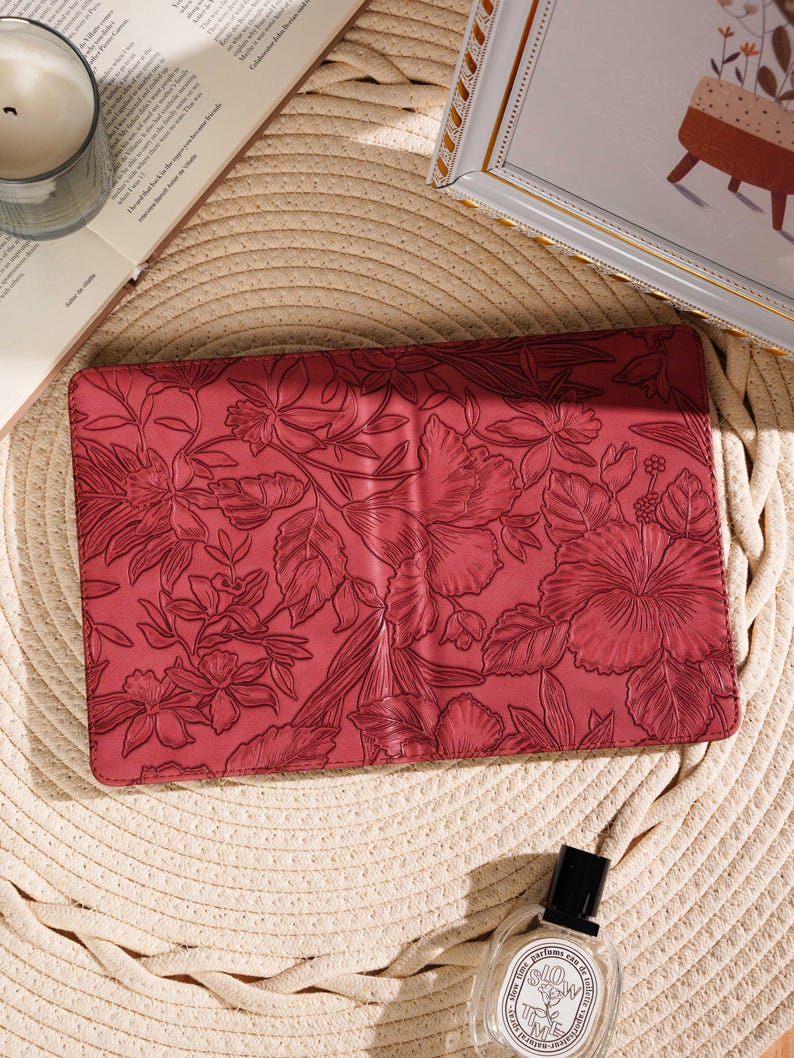 May include: A red floral-patterned clutch with a detailed embossed design. The clutch is rectangular and features a variety of flowers and leaves. The image is set on a straw mat with a candle, a book, and a perfume bottle.