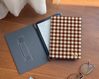 Puede incluir: Una funda de lector electrónico gris abierta con un soporte, junto a una funda de lector electrónico cerrada con un patrón de cuadros marrón y crema. La funda tiene iniciales doradas. Un tubo de crema azul y gafas redondas también están sobre la superficie de madera.