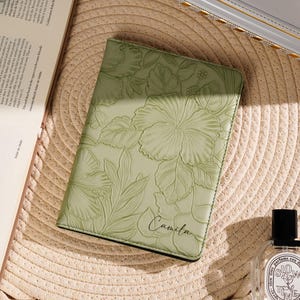 May include: A light green leather journal with embossed floral designs, including hibiscus flowers and leaves. The name "Camila" is written in script. The journal is on a straw hat, with a book and perfume bottle nearby.