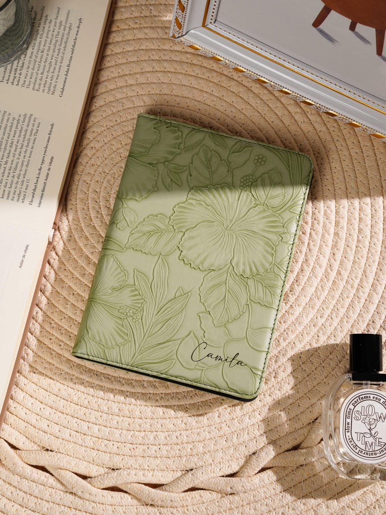 May include: A light green leather journal with embossed floral designs, including hibiscus flowers and leaves. The name "Camila" is written in script. The journal is on a straw hat, with a book and perfume bottle nearby.