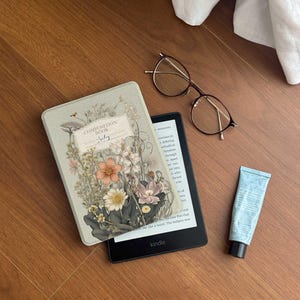 以下が含まれることがあります： 画面の下部に白い文字で「kindle」と書かれた白黒のKindle電子書籍リーダー。画面には本のテキストが表示されています。花柄のデザインと「Composition Book」と「Jatay」の文字が書かれたコンポジションノートが電子書籍リーダーの上に置かれています。木製の表面に眼鏡と青いハンドクリームのチューブが置かれています。