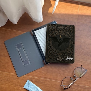 May include: An open grey e-reader case with a stand, a black and gold decorative e-reader cover with a dragon design, a tube of blue cream, and a pair of glasses on a wooden surface.
