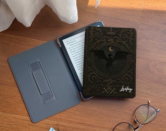 May include: An open grey e-reader case with a stand, a black and gold decorative e-reader cover with a dragon design, a tube of blue cream, and a pair of glasses on a wooden surface.