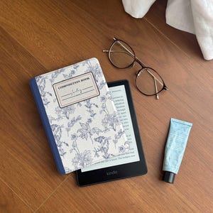Puede incluir: Un lector de libros electrónicos Kindle con una funda con estampado floral, un par de gafas y un tubo de loción azul están colocados sobre una superficie de madera. La funda dice "COMPOSITION BOOK" y "Jockey".