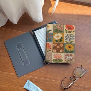 May include: An e-reader device in a protective case featuring a grid of colorful floral illustrations, including poppies, daisies, and sunflowers. The case has a gray interior with a hand strap. Nearby are round-framed glasses and a small blue tube.