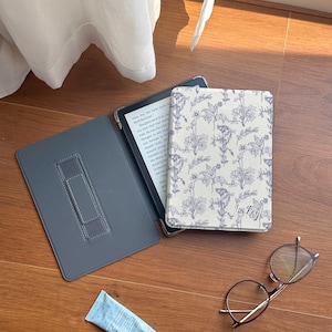 Op de afbeelding: Een grijze e-reader met een hoes met een bloemenpatroon. De hoes heeft de tekst "Lucky" erop. Er zijn ook een bril en een tube handcrème op een houten oppervlak.