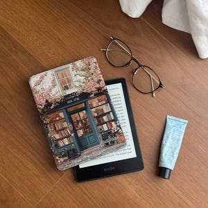 Könnte beinhalten: Ein schwarzer Kindle E-Reader mit einem floralen Buchcover, das ein Buchgeschäft mit dem Text "BOOK STORE" zeigt. Das Cover hat rosa Blumen und eine blaue Tür. Auf dem Bild sind auch eine Brille und eine blaue Tube Handcreme zu sehen.