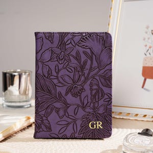 May include: A purple leather passport holder with a floral embossed design. The cover features a detailed pattern of flowers and leaves. Gold initials "GR" are visible on the lower right corner. The passport holder is on a cream-colored surface.