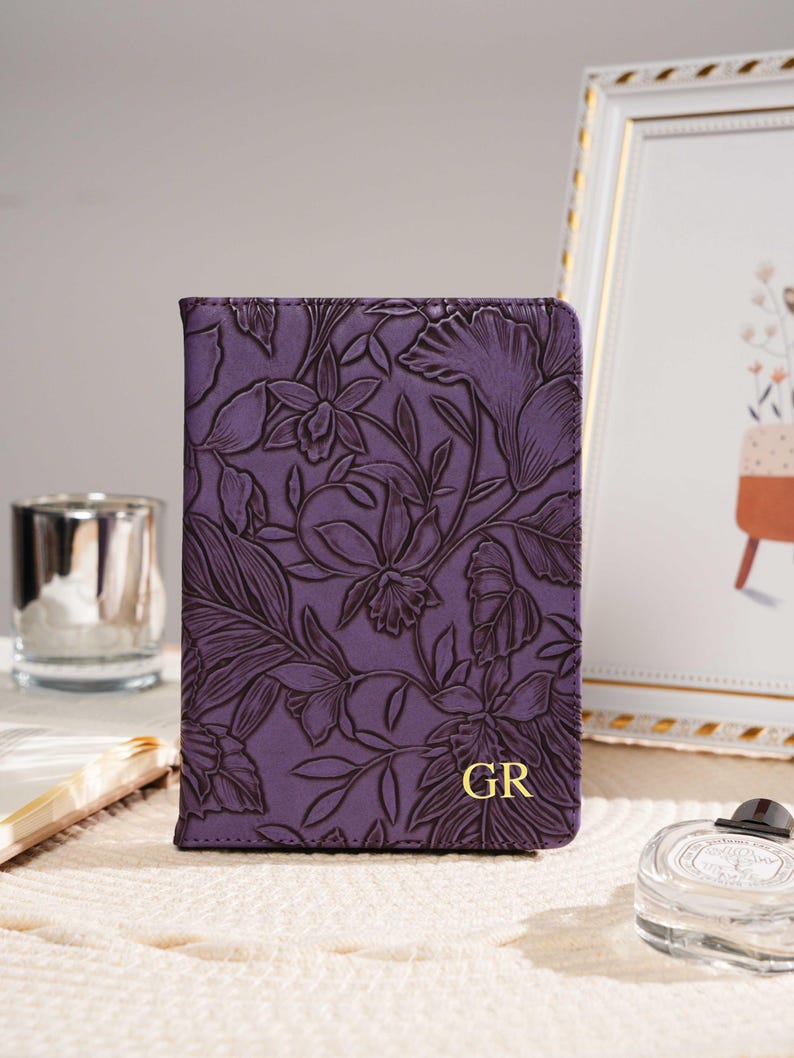 May include: A purple leather passport holder with a floral embossed design. The cover features a detailed pattern of flowers and leaves. Gold initials "GR" are visible on the lower right corner. The passport holder is on a cream-colored surface.