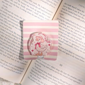 Puede incluir: Un pin de acr&iacute;lico transparente con un pato de dibujos animados con sombrero y bufanda rosas, con el texto "Silly Goose on the Loose". El pin est&aacute; sobre un fondo de rayas rosas y blancas.