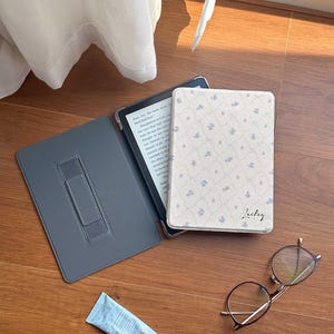 May include: An e-reader device in a cream-colored case with a blue floral pattern and the name Luckey printed in black script. The case is open, revealing a dark gray interior with a hand strap, alongside a pair of round glasses and a blue tube.