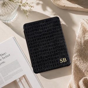 May include: A black e-reader case with a textured crocodile skin pattern and the gold-colored initials SB embossed in the bottom right corner. The rectangular case features a sleek, structured design with a consistent grid-like embossed finish.