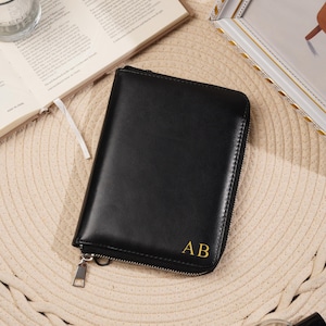 Könnte beinhalten: Ein schwarzes, rechteckiges Organizer-Etui mit Reißverschluss und glatter Oberfläche. In der unteren rechten Ecke befinden sich die goldfarben geprägten Initialen AB. Das Etui verfügt über einen silberfarbenen Metall-Reißverschluss.