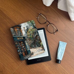 Könnte beinhalten: Ein schwarzer Kindle-E-Reader mit einer Hülle, die ein Aquarellgemälde eines Buchladens zeigt. Das Bild enthält auch eine runde Brille, eine Tube blaue Lotion und ein weißes Tuch auf einer Holzoberfläche.