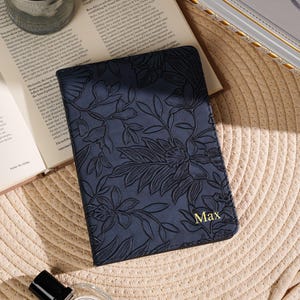 May include: A navy blue passport holder with a floral embossed design and the name "Max" in gold lettering. The holder is on a straw mat, next to a candle, a book, and a perfume bottle.