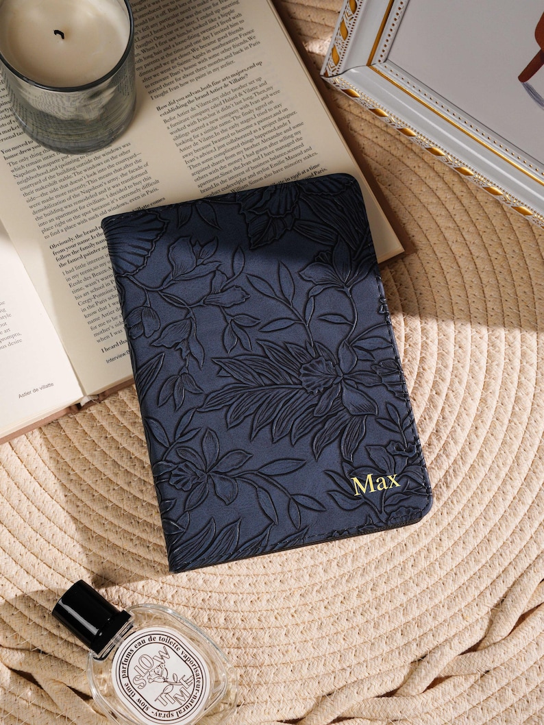 May include: A navy blue passport holder with a floral embossed design and the name "Max" in gold lettering. The holder is on a straw mat, next to a candle, a book, and a perfume bottle.