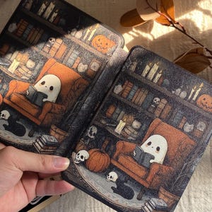 K&ouml;nnte beinhalten: Zwei dekorative Buchh&uuml;llen mit Halloween-Motiv. Jede H&uuml;lle zeigt einen Geist, der in einem gem&uuml;tlichen Sessel vor einem B&uuml;cherregal liest. K&uuml;rbisse, Totenk&ouml;pfe und Kerzen tragen zur gruseligen Atmosph&auml;re bei. Die H&uuml;llen werden in einer Hand gehalten.