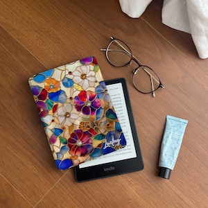 Puede incluir: Un lector de libros electrónicos Kindle con una funda floral colorida, gafas marrones y un tubo de loción azul descansan sobre una superficie de madera. La funda presenta un diseño de vidriera con flores rojas, azules y moradas.