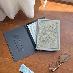 Op de afbeelding: Een saliegroene e-readerhoes met een symmetrisch bloemenpatroon in blauw en wit, een grijze binnenkant met een handriem, een kleine lichtblauwe tube crème en een ronde bril met een dun, donker metalen montuur.