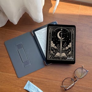 May include: An e-reader device featuring a black protective cover with a white graphic illustration of a sword, an open book, and a crescent moon. The device is displayed alongside a gray folding case, a small tube of cream, and round glasses.
