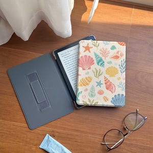 May include: An e-reader device in a protective case with a cream-colored cover featuring a colorful pattern of seashells, starfish, and coral. The case interior is dark gray with an elastic hand strap. Nearby are round glasses and a small blue tube.