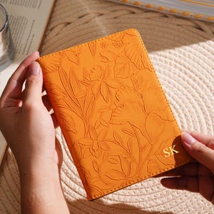 May include: Orange leather passport holder with embossed floral design. The initials "SK" are in gold on the lower right corner. The passport holder is held by two hands.
