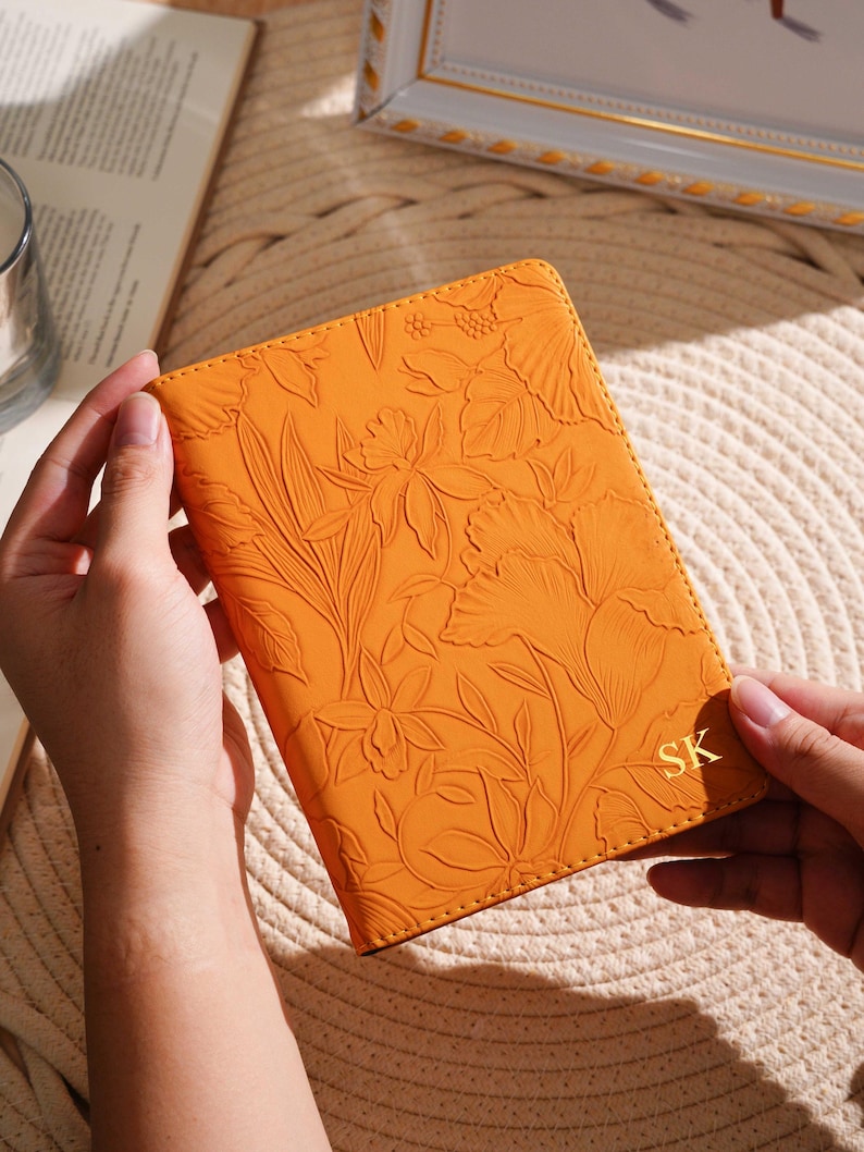 May include: Orange leather passport holder with embossed floral design. The initials "SK" are in gold on the lower right corner. The passport holder is held by two hands.