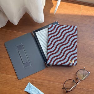 Puede incluir: Una funda de lector electrónico gris abierta con un soporte, una cubierta estampada con un diseño ondulado granate y azul claro, y un tubo de crema azul están sobre una superficie de madera. También hay gafas con montura dorada.