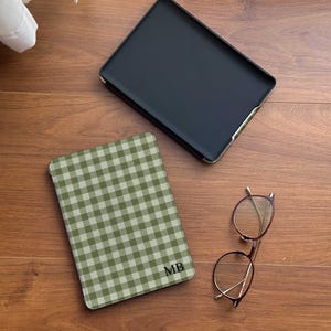 Puede incluir: Una funda negra para tableta con ribete dorado se encuentra junto a una funda para tableta a cuadros verdes y blancos con las iniciales "MB". Un par de gafas redondas con montura dorada descansa cerca. Los art&iacute;culos est&aacute;n sobre una superficie de madera.