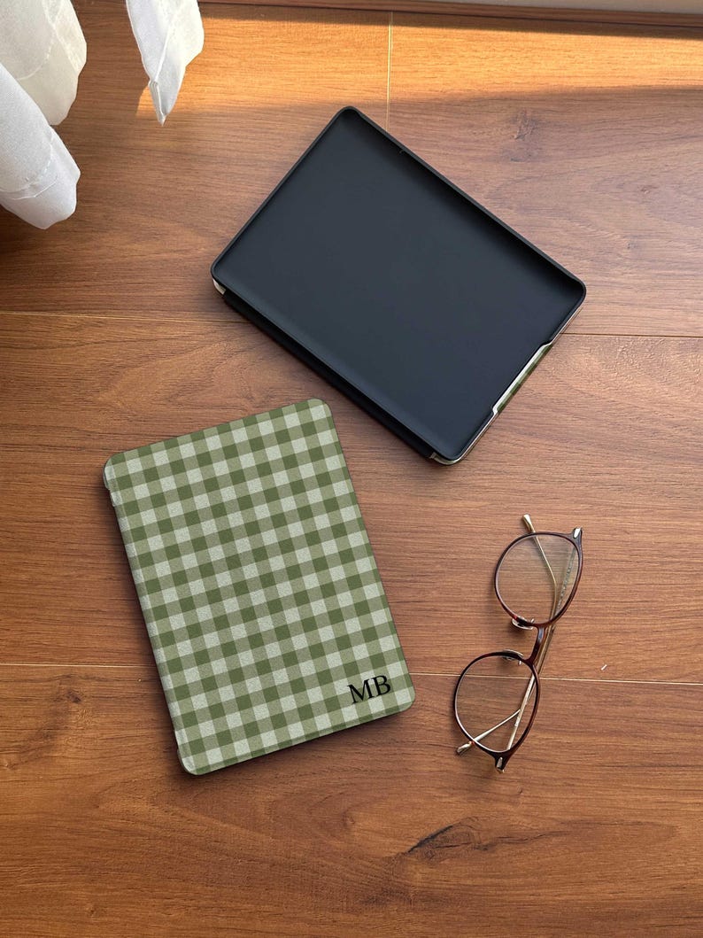 Puede incluir: Una funda negra para tableta con ribete dorado se encuentra junto a una funda para tableta a cuadros verdes y blancos con las iniciales "MB". Un par de gafas redondas con montura dorada descansa cerca. Los art&iacute;culos est&aacute;n sobre una superficie de madera.