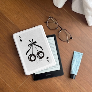 以下が含まれることがあります： 黒いチェリーのデザインと「Luckey」の文字が入った白いカバーのKindle電子書籍リーダー。メガネ、ローションチューブ、白い布も木製の表面に写っています。