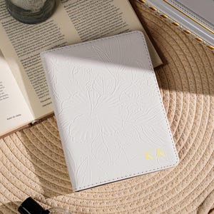 May include: White passport holder with embossed floral design and gold initials "KK". The cover is on a woven straw surface, next to an open book, a candle in a glass, and a perfume bottle.
