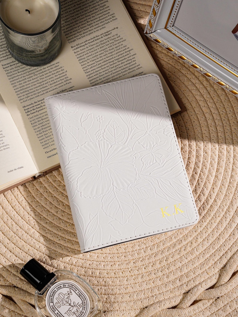 May include: White passport holder with embossed floral design and gold initials "KK". The cover is on a woven straw surface, next to an open book, a candle in a glass, and a perfume bottle.
