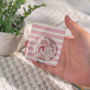 Puede incluir: Un soporte transparente para tel&eacute;fono con un dibujo de un ganso de dibujos animados con un sombrero de vaquero y una bufanda roja, con el texto "Silly Goose on the Loose". El soporte est&aacute; sobre un respaldo a rayas rosas y blancas.