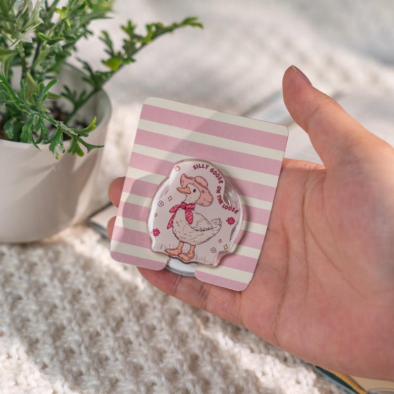 Puede incluir: Un soporte transparente para tel&eacute;fono con un dibujo de un ganso de dibujos animados con un sombrero de vaquero y una bufanda roja, con el texto "Silly Goose on the Loose". El soporte est&aacute; sobre un respaldo a rayas rosas y blancas.
