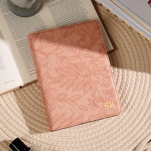 May include: A blush-colored leather journal with an embossed floral design and gold initials "SB". The journal is open on a book, with a candle and perfume bottle nearby. The background is a woven straw mat.
