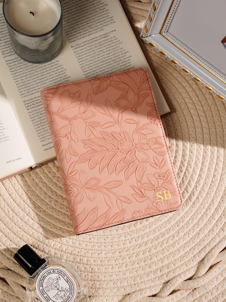 May include: A blush-colored leather journal with an embossed floral design and gold initials "SB". The journal is open on a book, with a candle and perfume bottle nearby. The background is a woven straw mat.