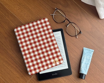Puede incluir: Una funda Kindle a cuadros rojos y blancos con iniciales doradas, un Kindle negro, gafas redondas y un tubo de loción azul sobre una superficie de madera.