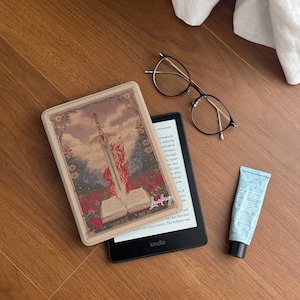 May include: A Kindle e-reader with a decorative case featuring a sword and open book design with flames. Also, a pair of glasses and a tube of blue lotion are on a wooden surface.